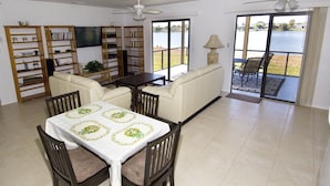 A spacious living room with a dining area, a ceiling fan, a TV, and a view of a lake through large sliding doors. - Lilsis by the Lake - Vacation Home (Port Charlotte)