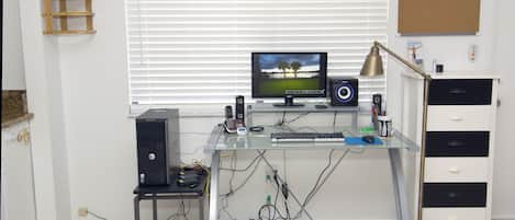 A home office with a desktop computer, monitor, keyboard, and speakers. There is a plant on the wall, a corkboard, and a desk lamp.