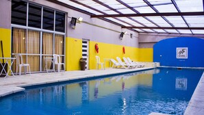 Una piscina interior con agua azul cristalina, sillas de descanso blancas y una pared amarilla y azul.