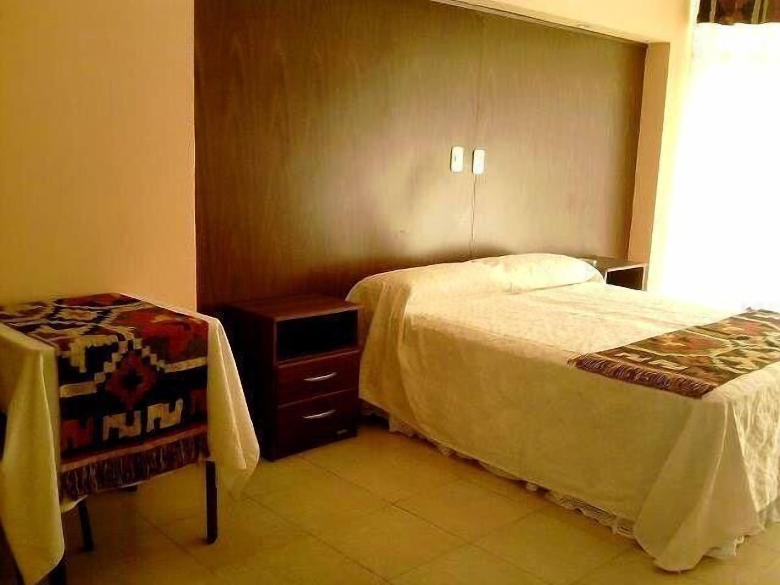 A bedroom with a bed, a nightstand, a small table with a patterned tablecloth, and a wardrobe.