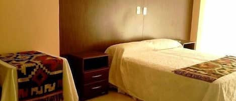 A bedroom with a bed, a nightstand, a small table with a patterned tablecloth, and a wardrobe.