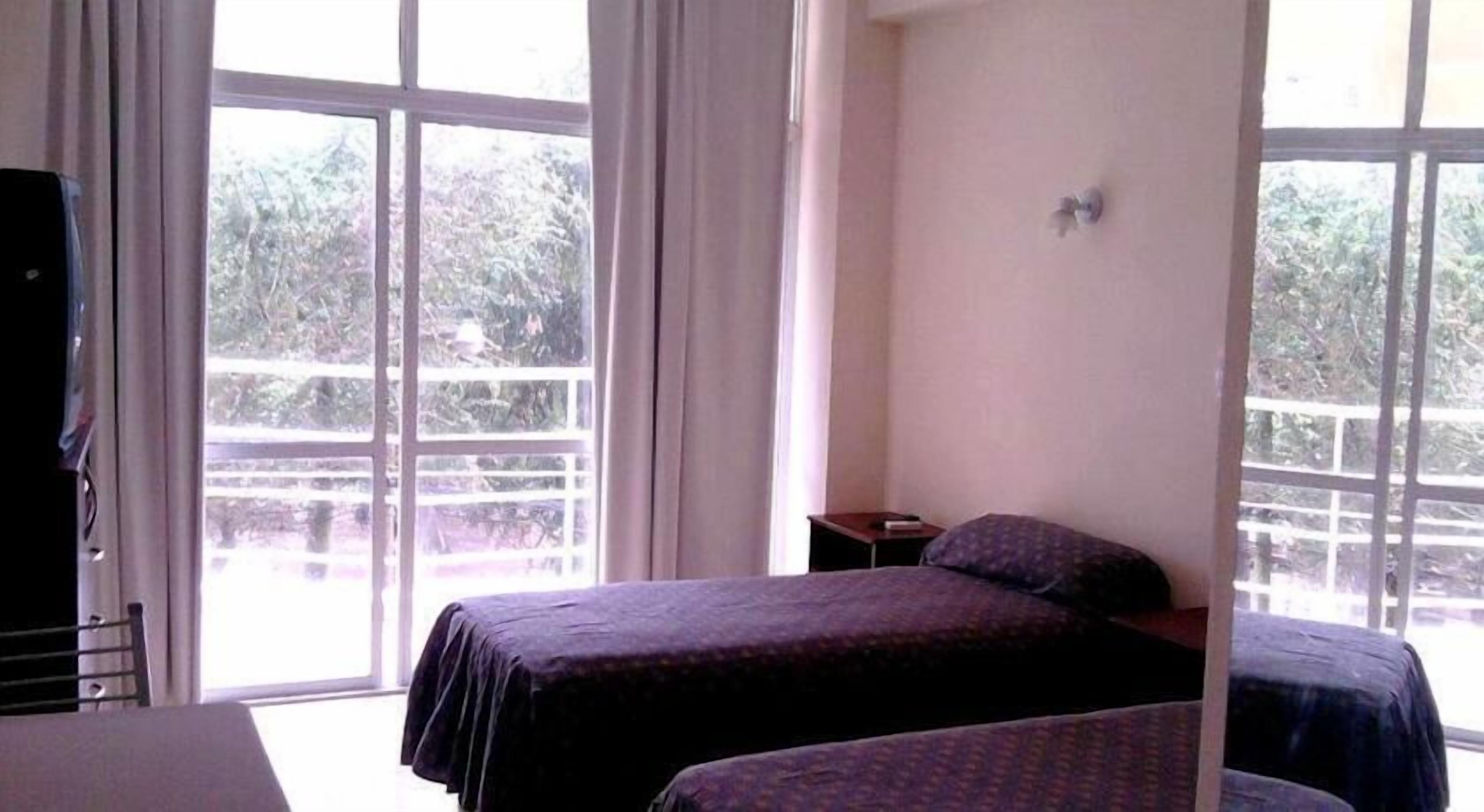 Ein Zimmer mit zwei Betten, großen Fenstern, einem kleinen Tisch und einer Wandleuchte.