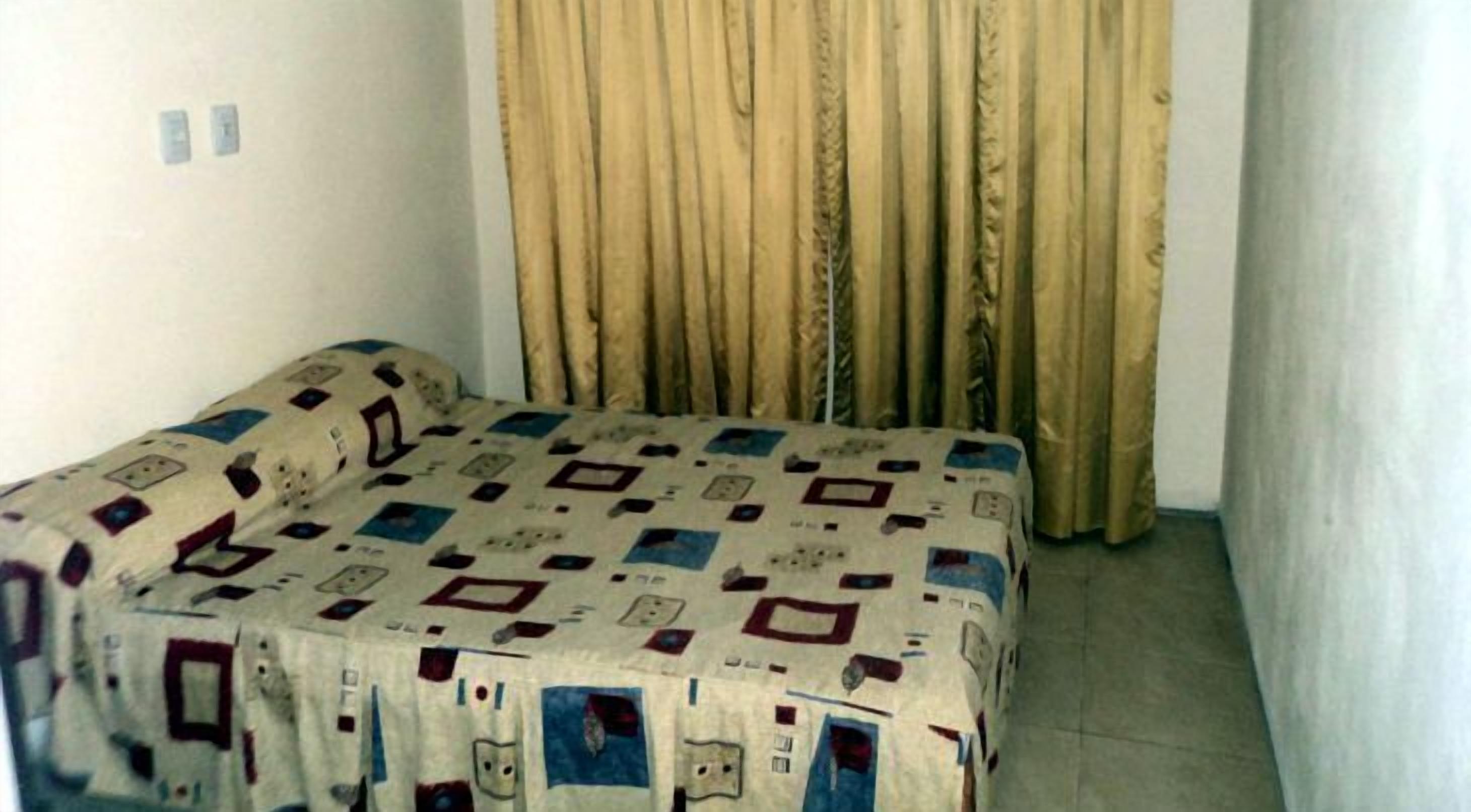 Una camera piccola con un letto singolo, una coperta a motivi, una tenda e un muro liscio.