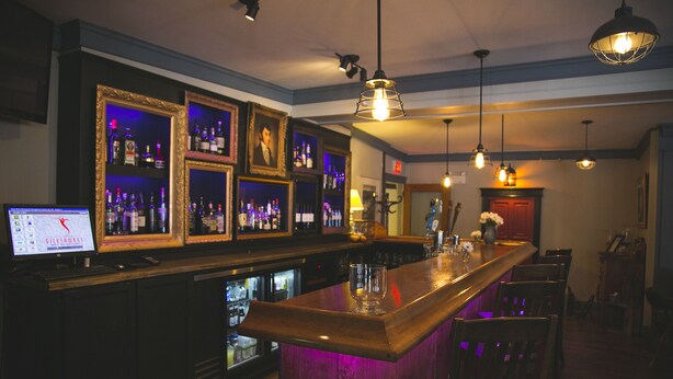 Een bar met een houten toonbank, planken met flessen, ingelijste foto's en hangende lampen.