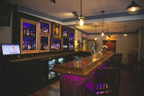 Uma área de bar com balcão de madeira, prateleiras com garrafas, quadros emoldurados e luminárias pendentes.