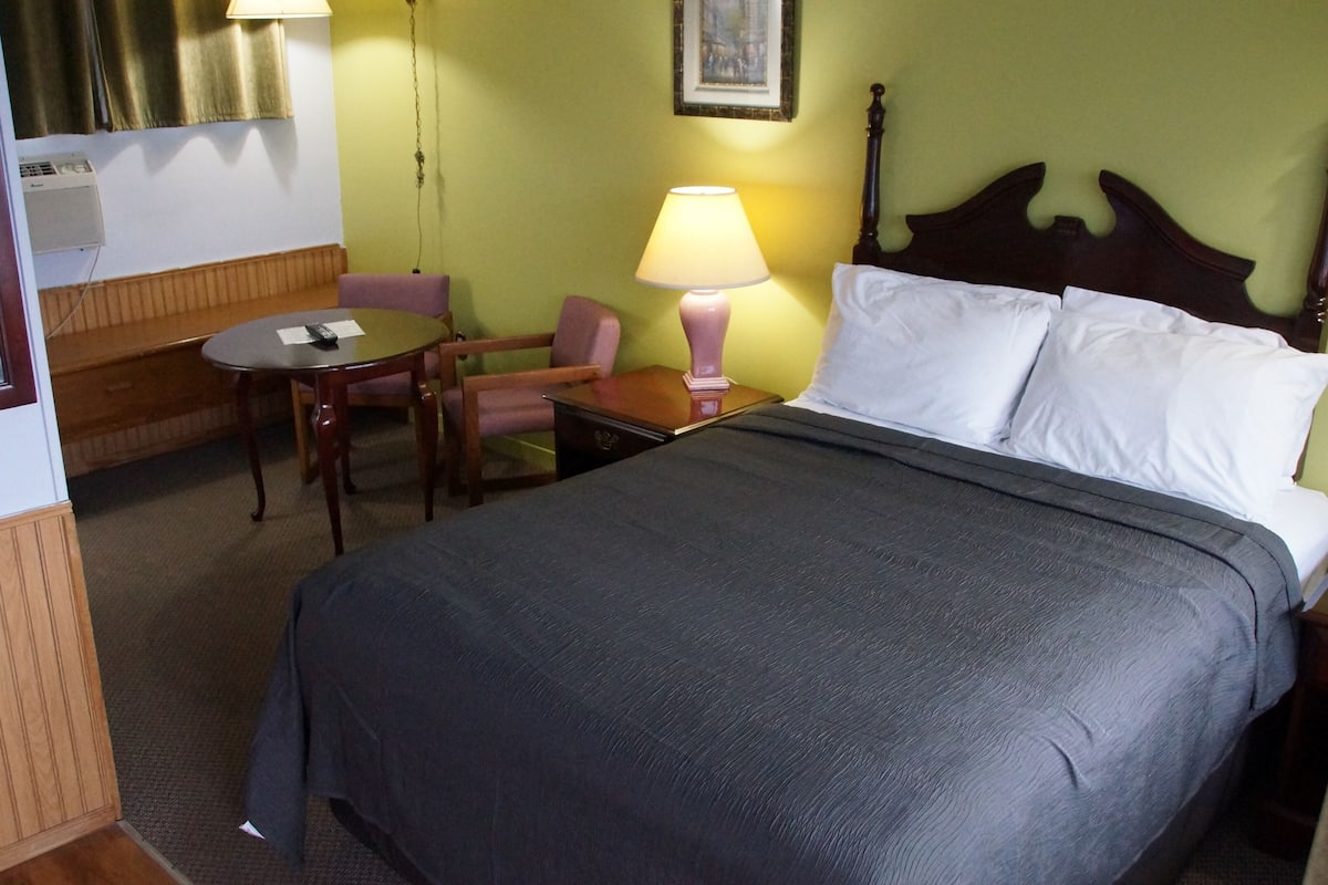 A hotel room with a bed, a desk with a lamp, a chair, and a small table.