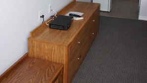 Habitación de hotel con un escritorio de madera, un televisor de pantalla plana montado en la pared, un espejo, un refrigerador blanco pequeño y una banca.