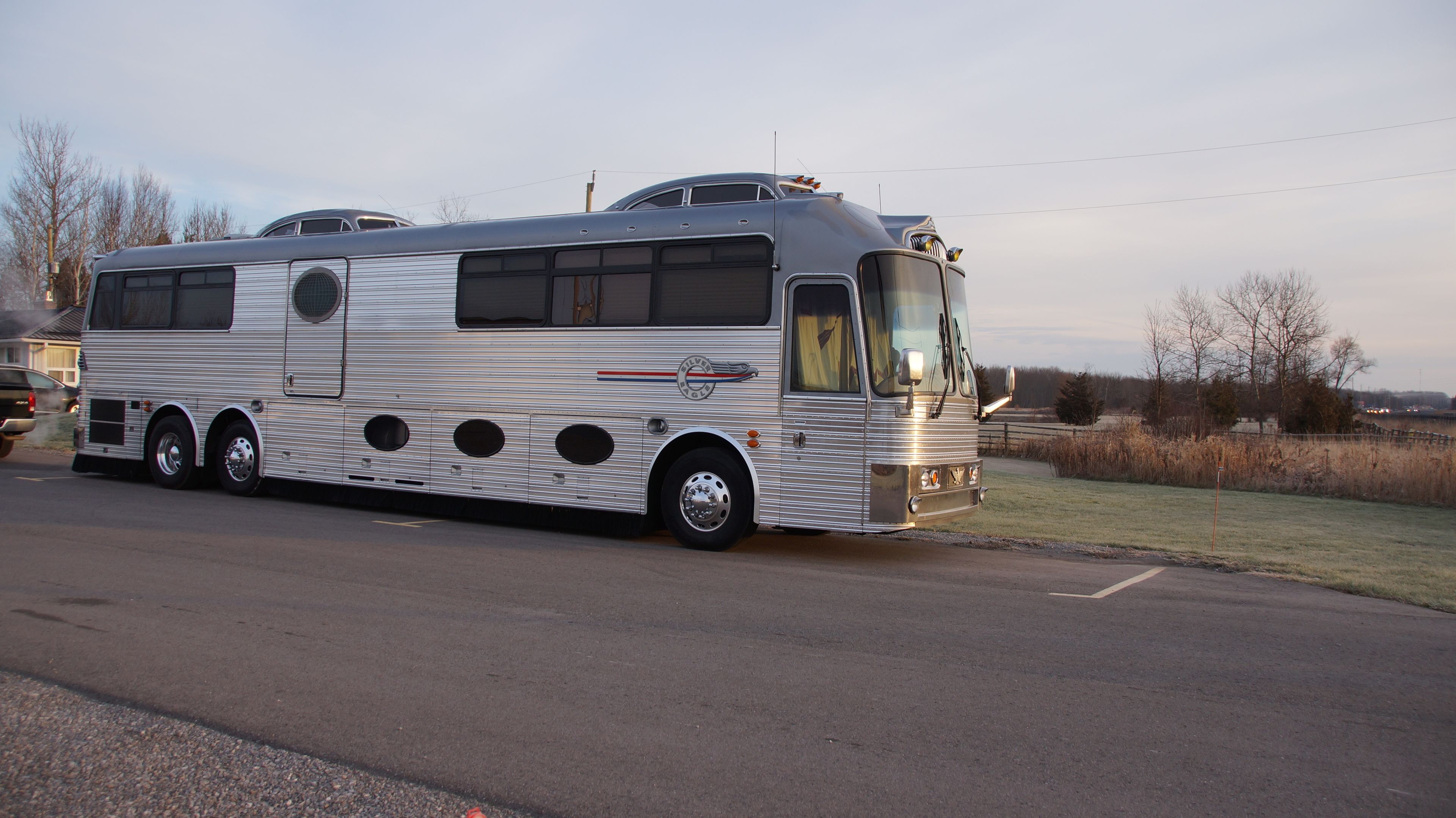 A large, silver bus with multiple windows and a front windshield, parked on a road with a grassy area and trees in the background.