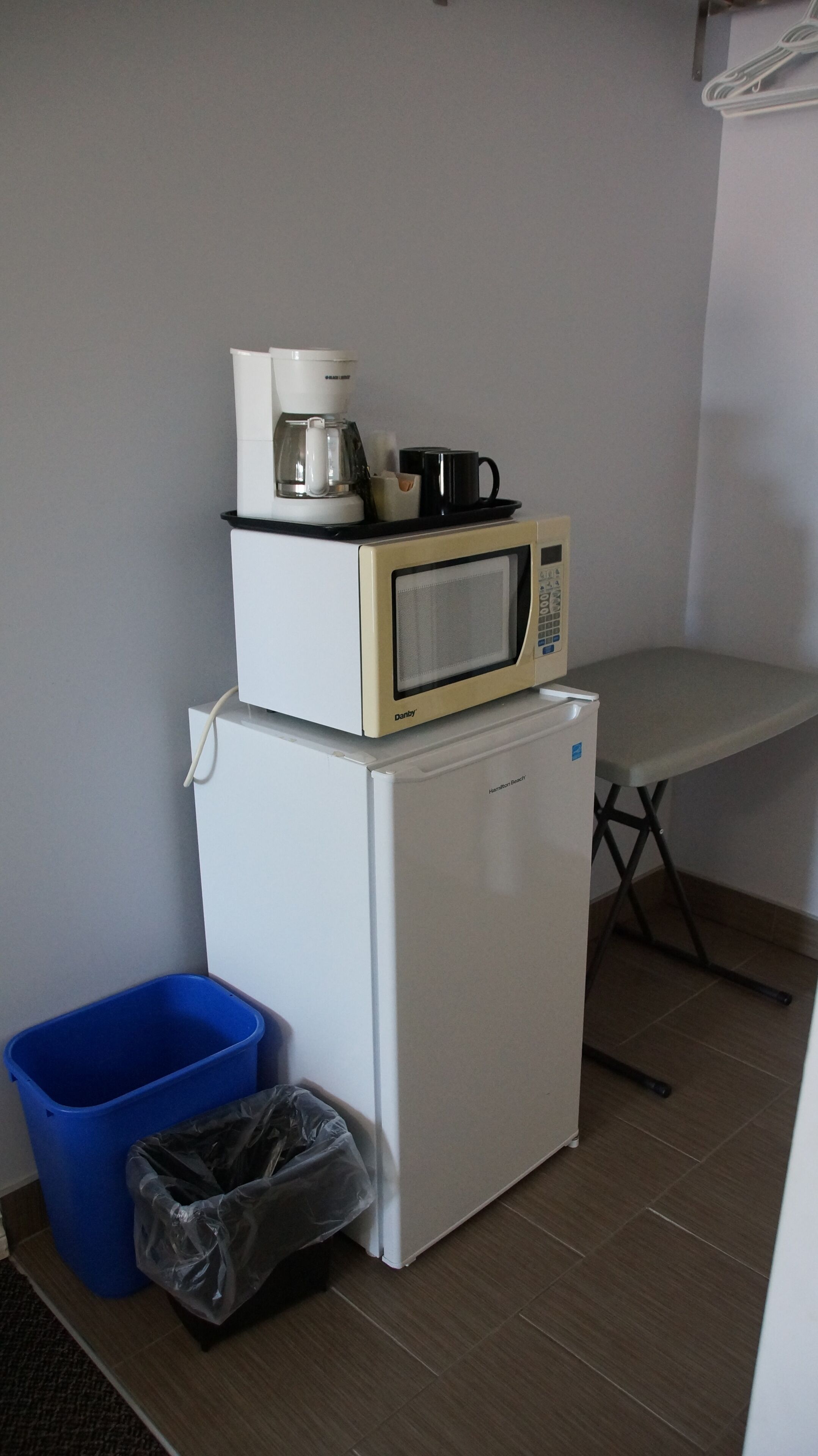 Ett litet, praktiskt kök med mikrovågsugn, kaffebryggare och ett litet kylskåp. En blå papperskorg och en plastpåse står på golvet.