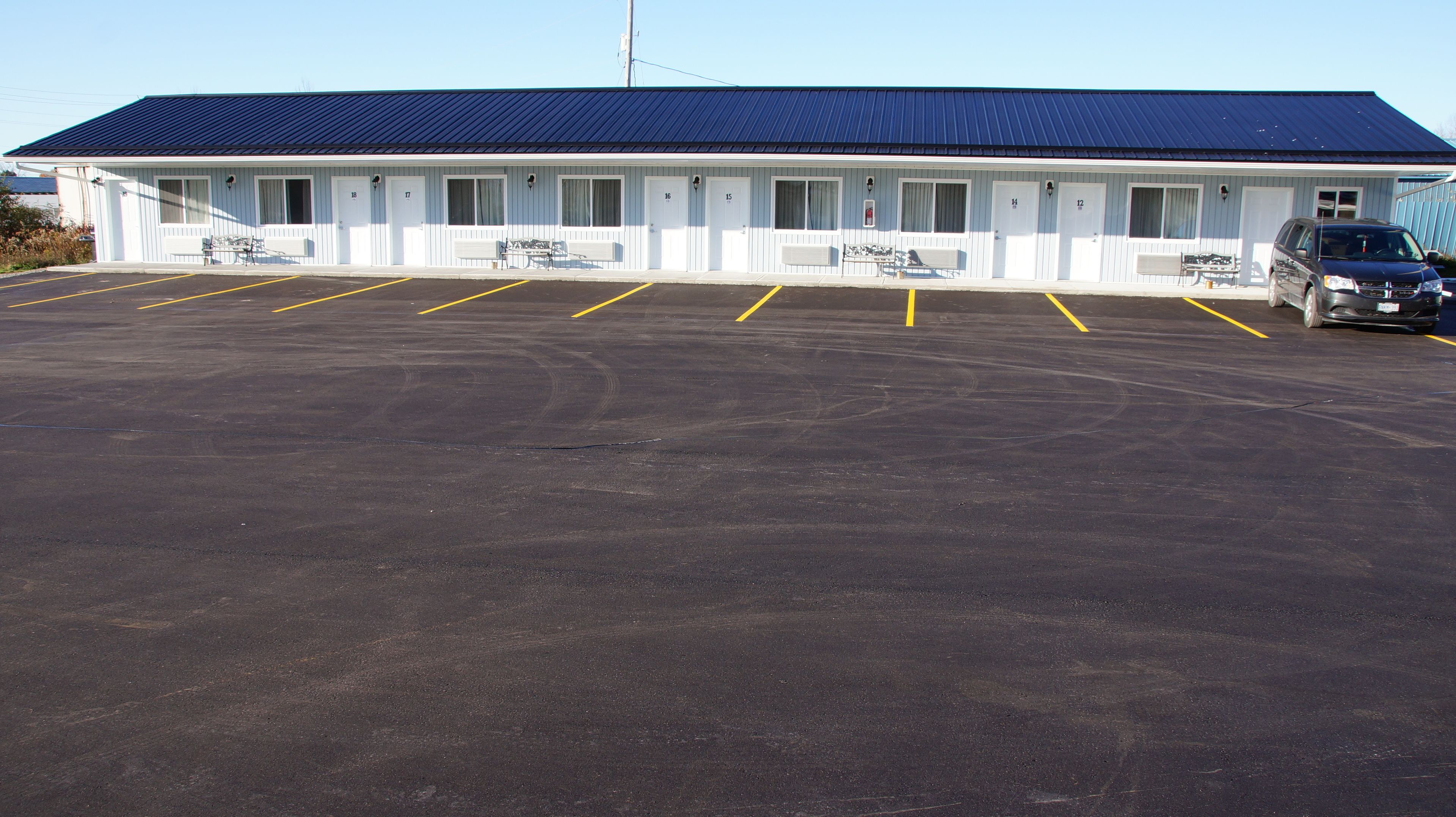 A motel with a blue roof and white exterior, featuring numbered doors from 12 to 18, and a parking lot with yellow lines.