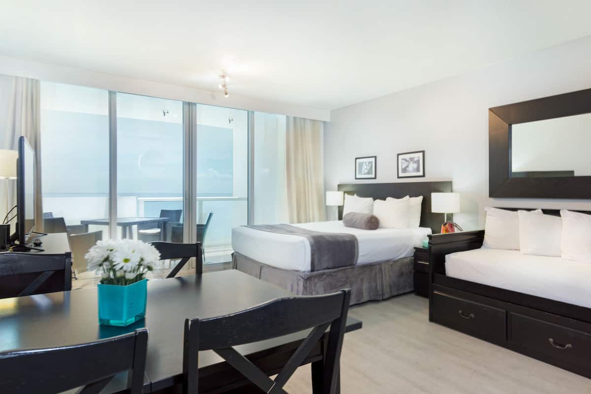 A hotel room with a large bed, a desk, a chair, a table with a vase of flowers, a balcony with a view of the sea, and a large mirror on the wall.