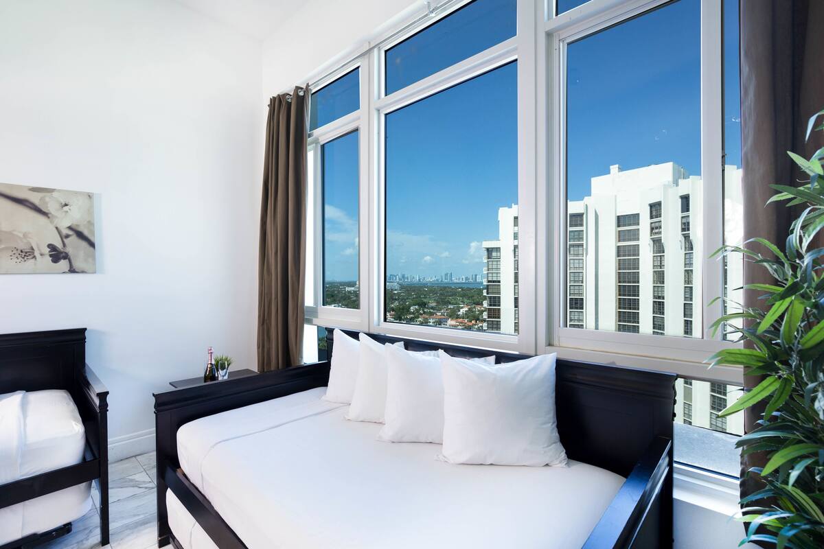 A hotel room with a large window, a bed with white bedding, a nightstand with a bottle, and a plant.