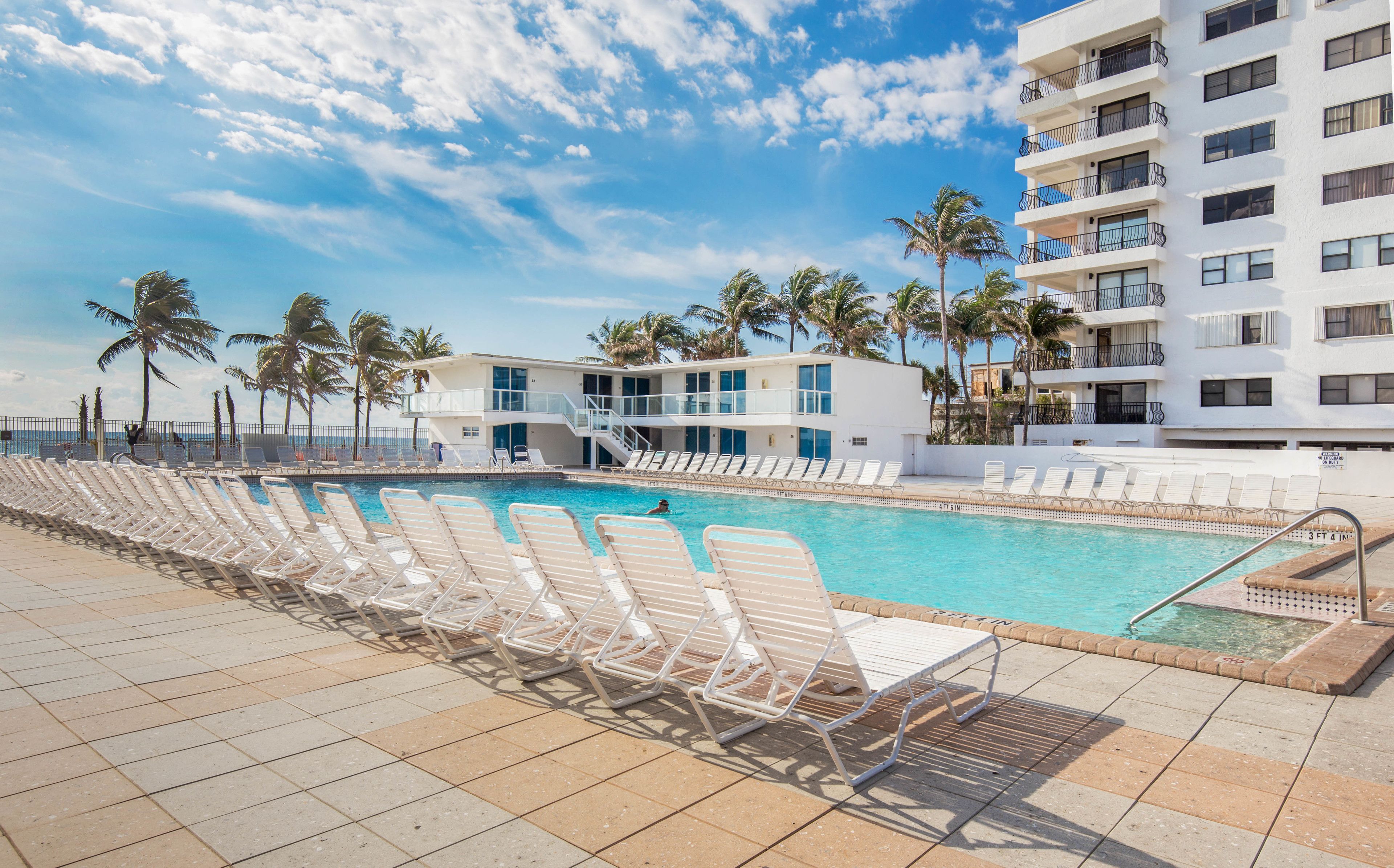 A poolside area with lounge chairs and a swimming pool, surrounded by palm trees and a building with balconies.