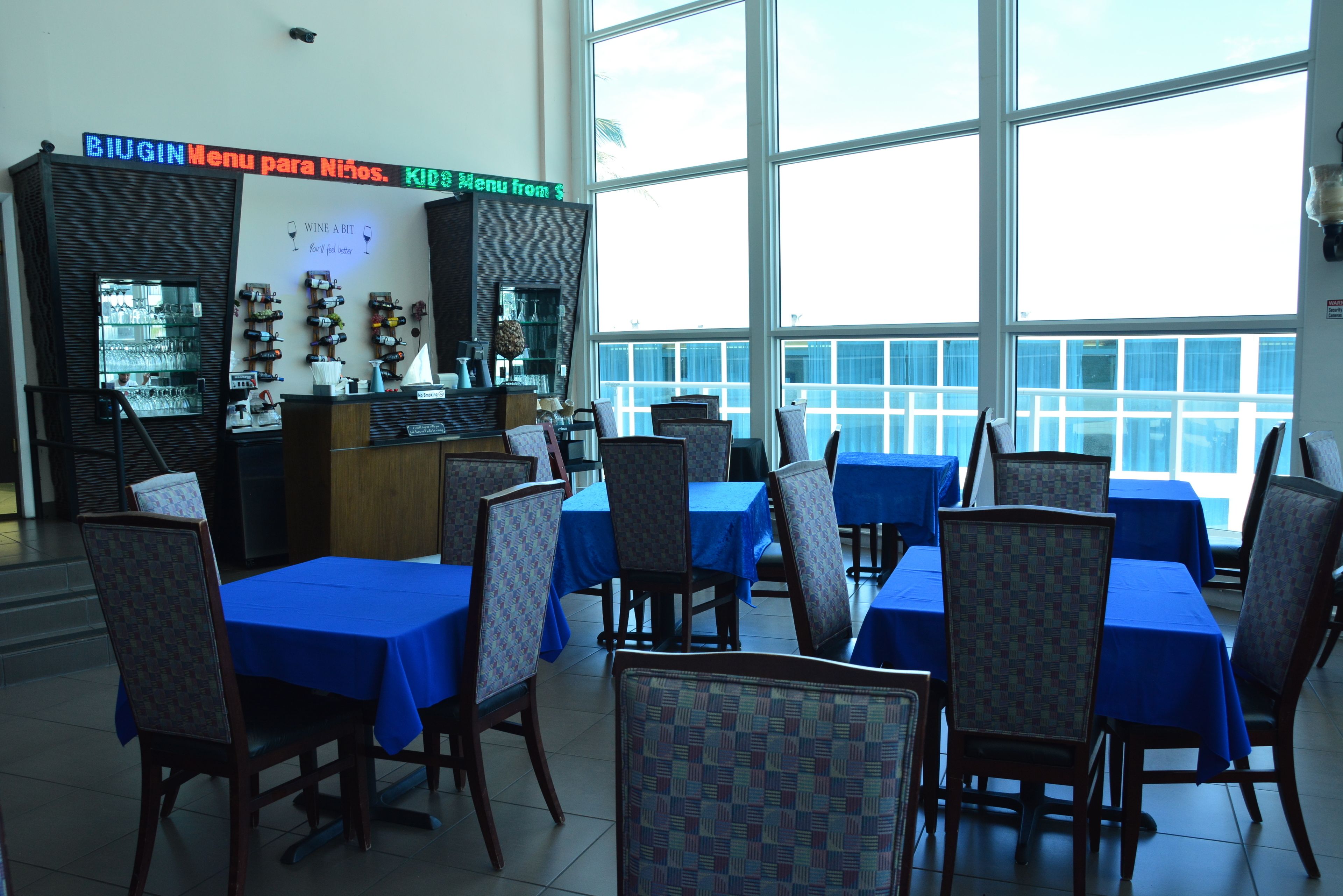 A restaurant with blue tablecloths and chairs, a bar area with wine glasses, and a view through large windows.
