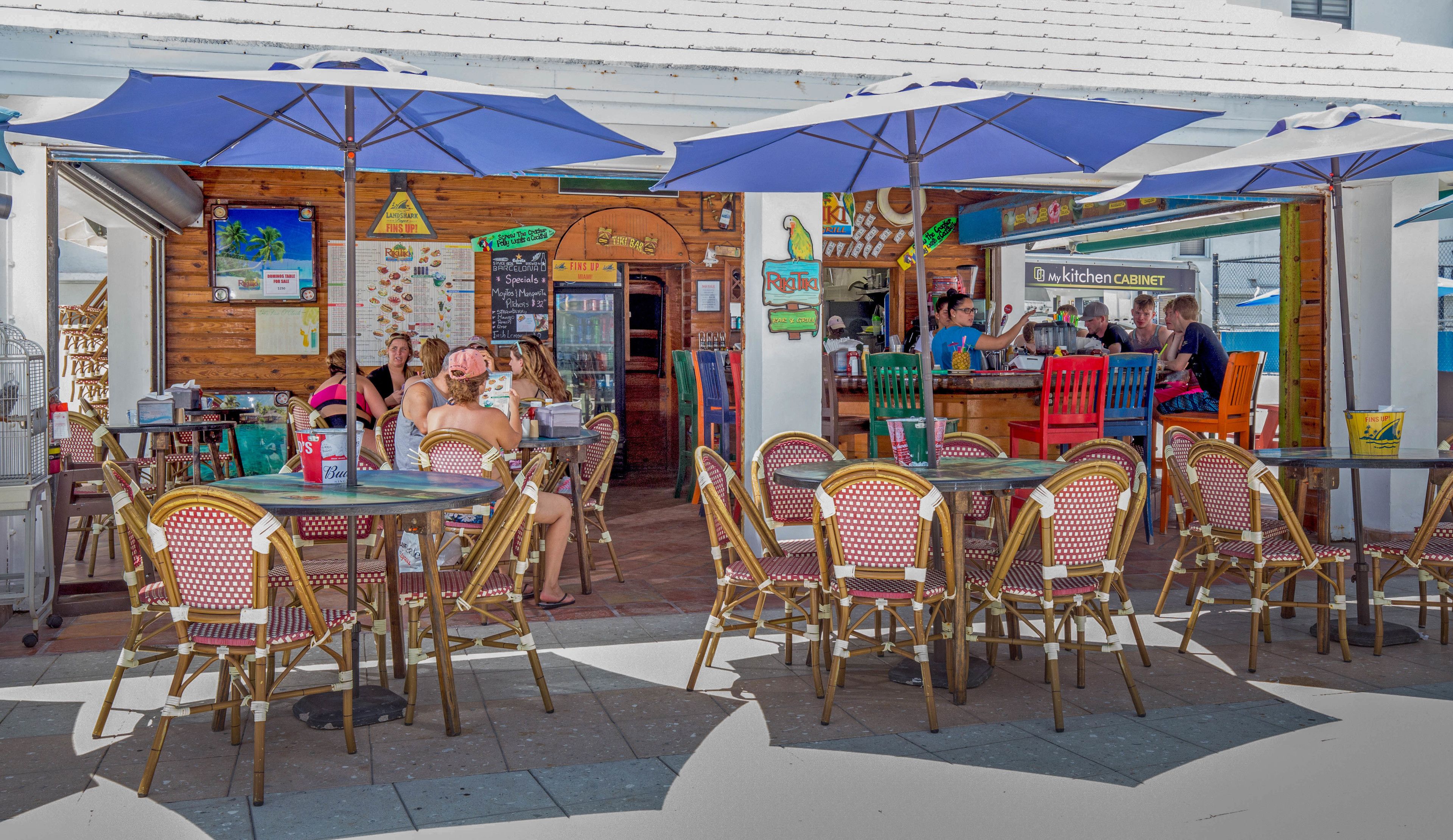An outdoor cafe with wicker tables and chairs, blue umbrellas, and a wooden facade.