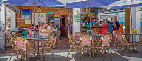 An outdoor cafe with wicker tables and chairs, blue umbrellas, and a wooden facade.