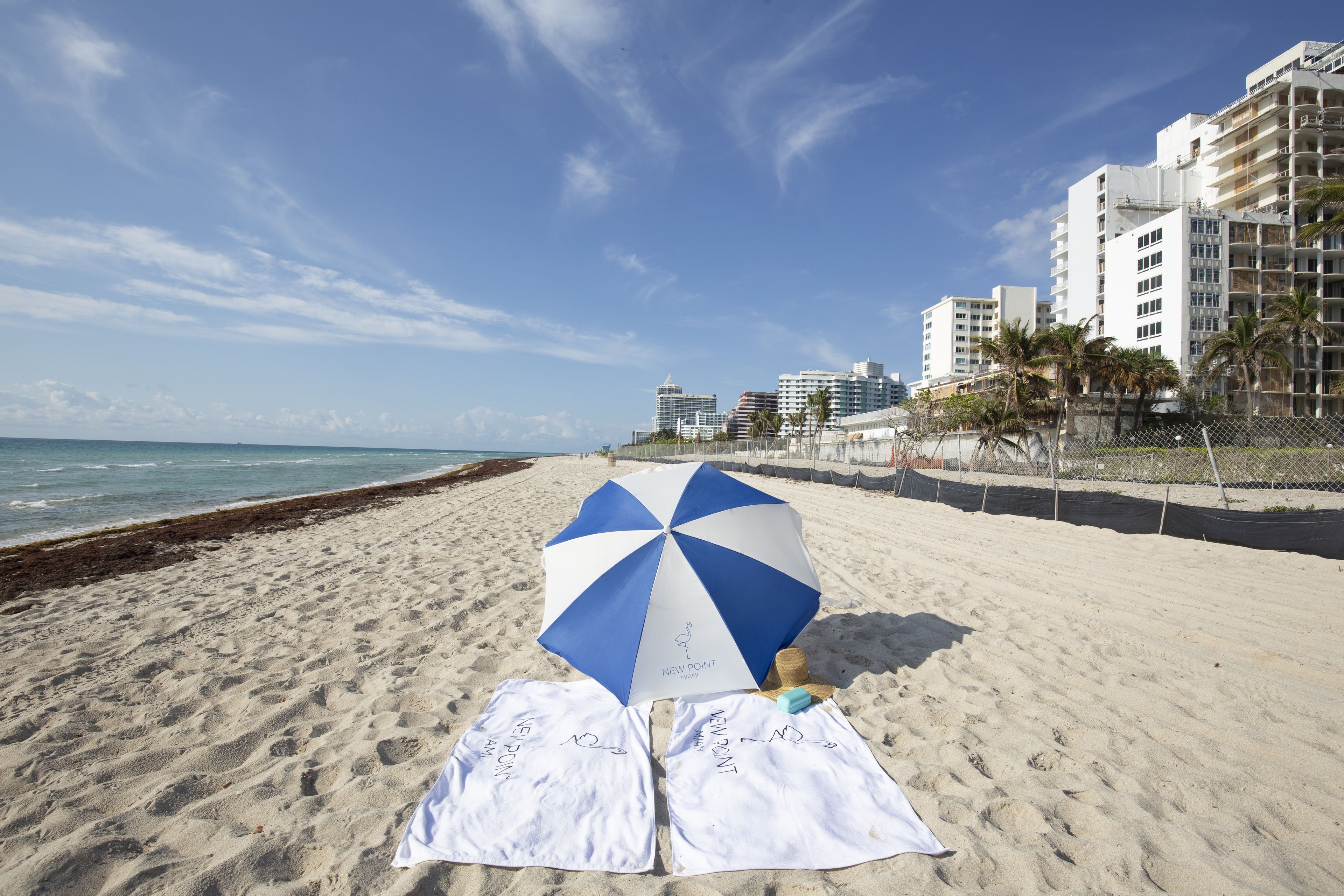 A beach with a blue and white umbrella, two white towels, and a fence.