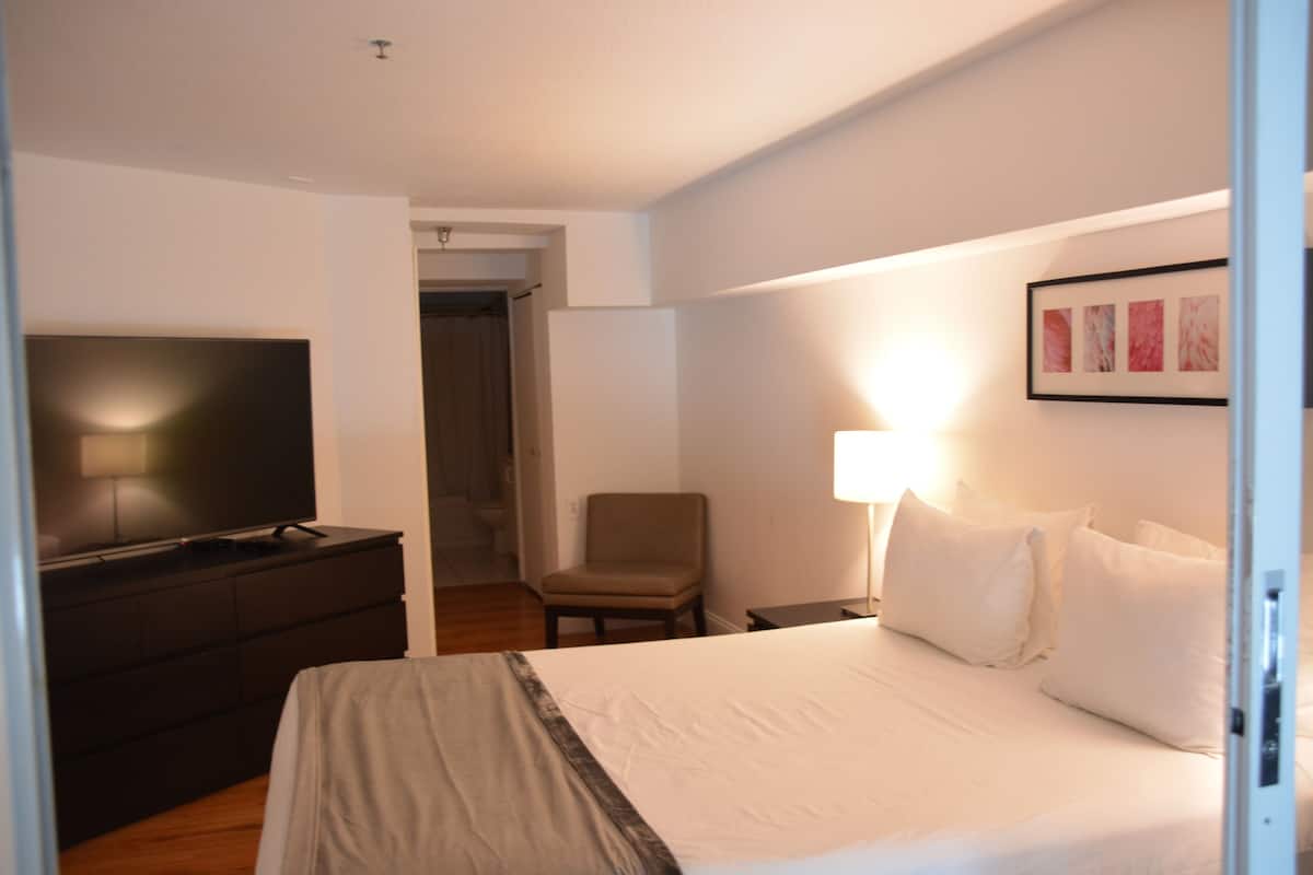 A hotel room with a bed, a TV on a dresser, a chair, and a nightstand with a lamp.