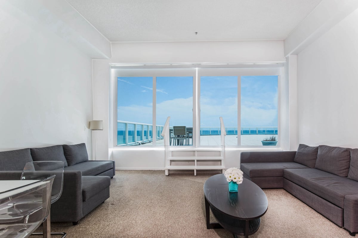 A modern living room with a large window overlooking a balcony with a view of the sea.