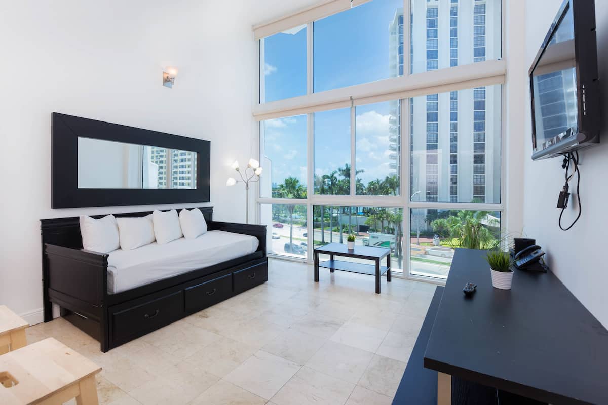 A modern hotel room with a large bed, a desk, a television, and a large window offering a city view.