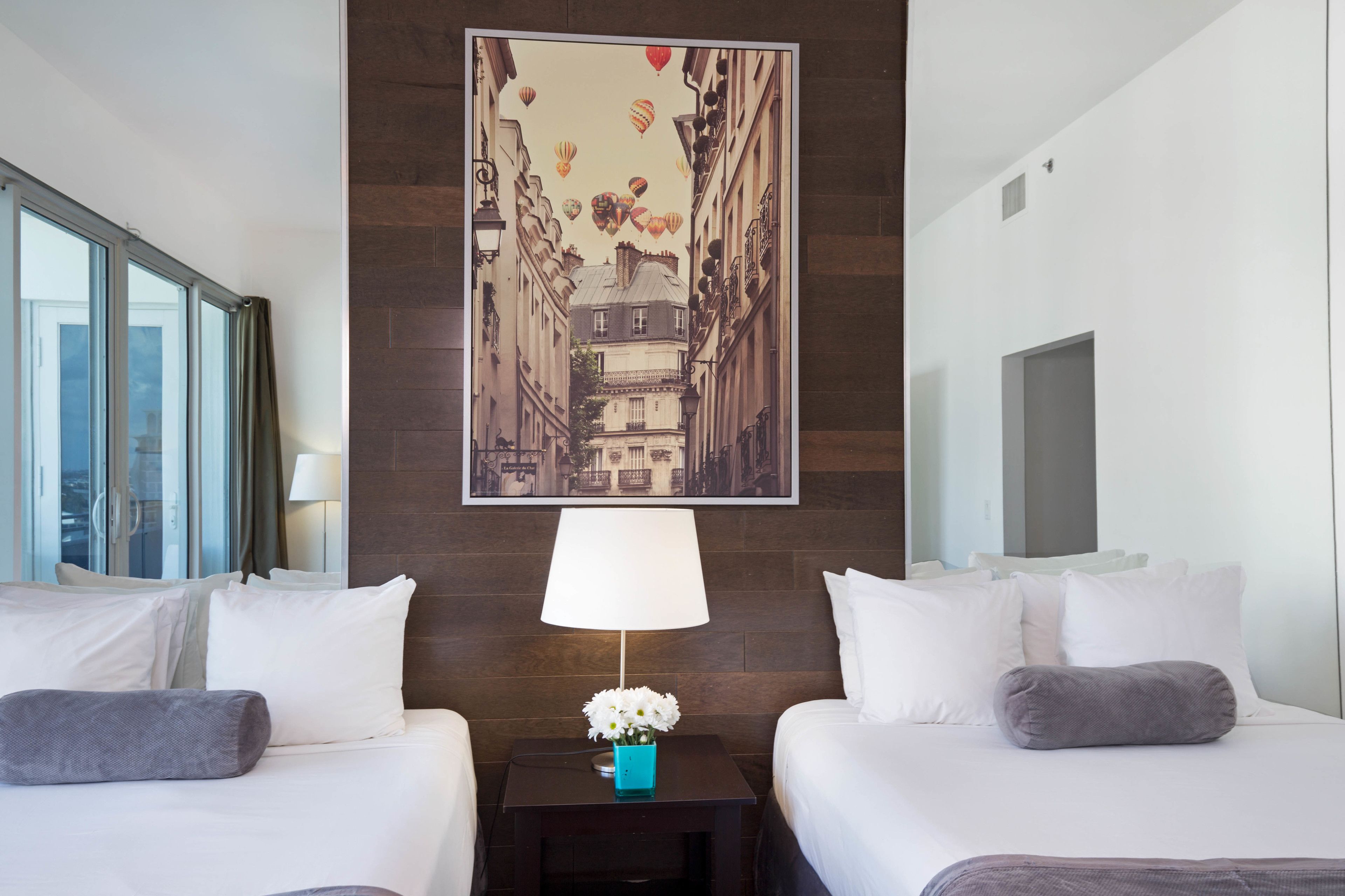 Une chambre d’hôtel avec deux lits, une table de chevet avec une lampe et des fleurs, un grand tableau encadré représentant une rue de la ville, et une fenêtre avec des rideaux.