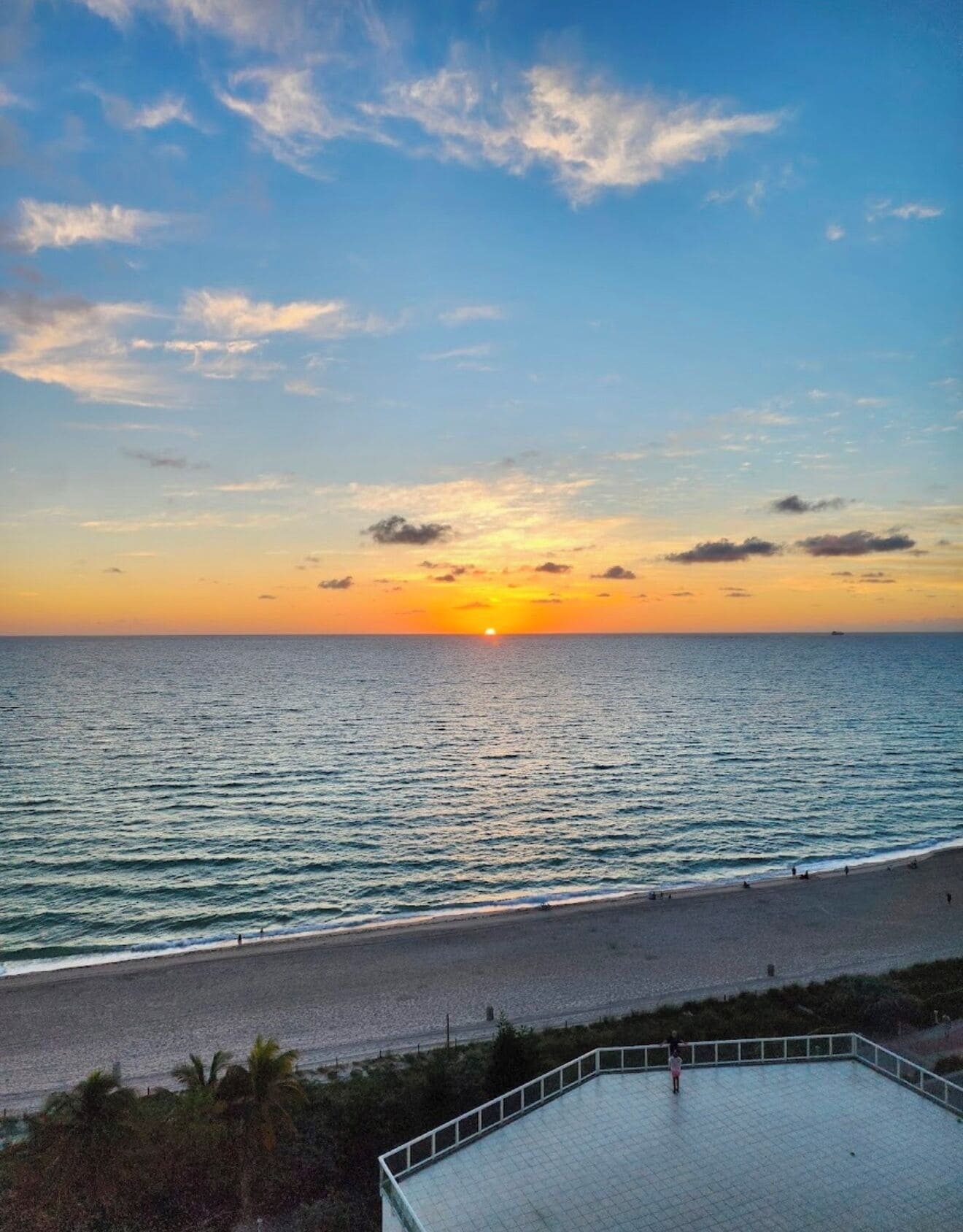 A beach with a sunset, calm sea, and a paved area with a railing.