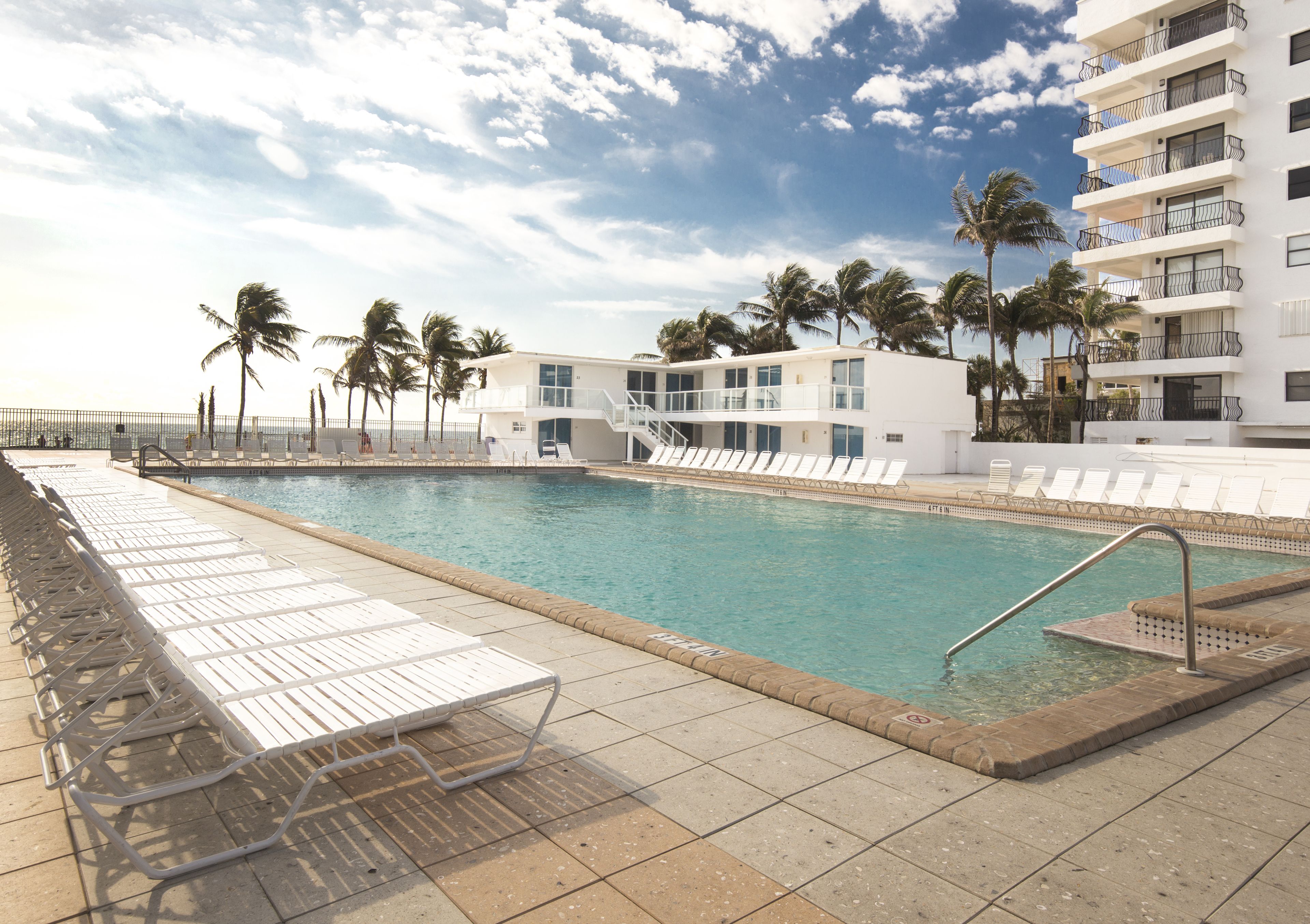 Zona de piscina con sillones de descanso, palmeras y un edificio moderno con balcones.