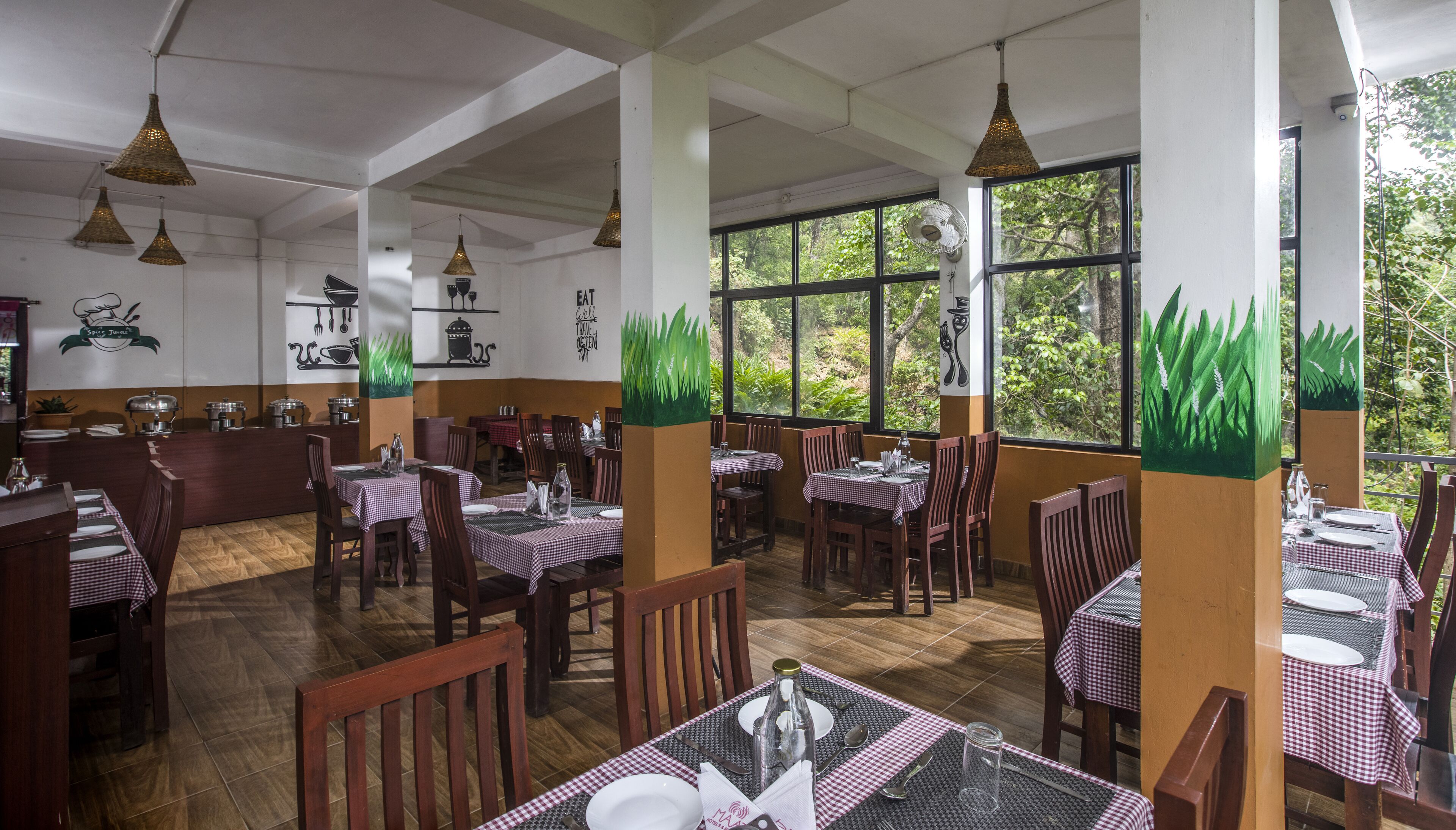 A restaurant with wooden tables and chairs, large windows with greenery outside, and decorative wall art.