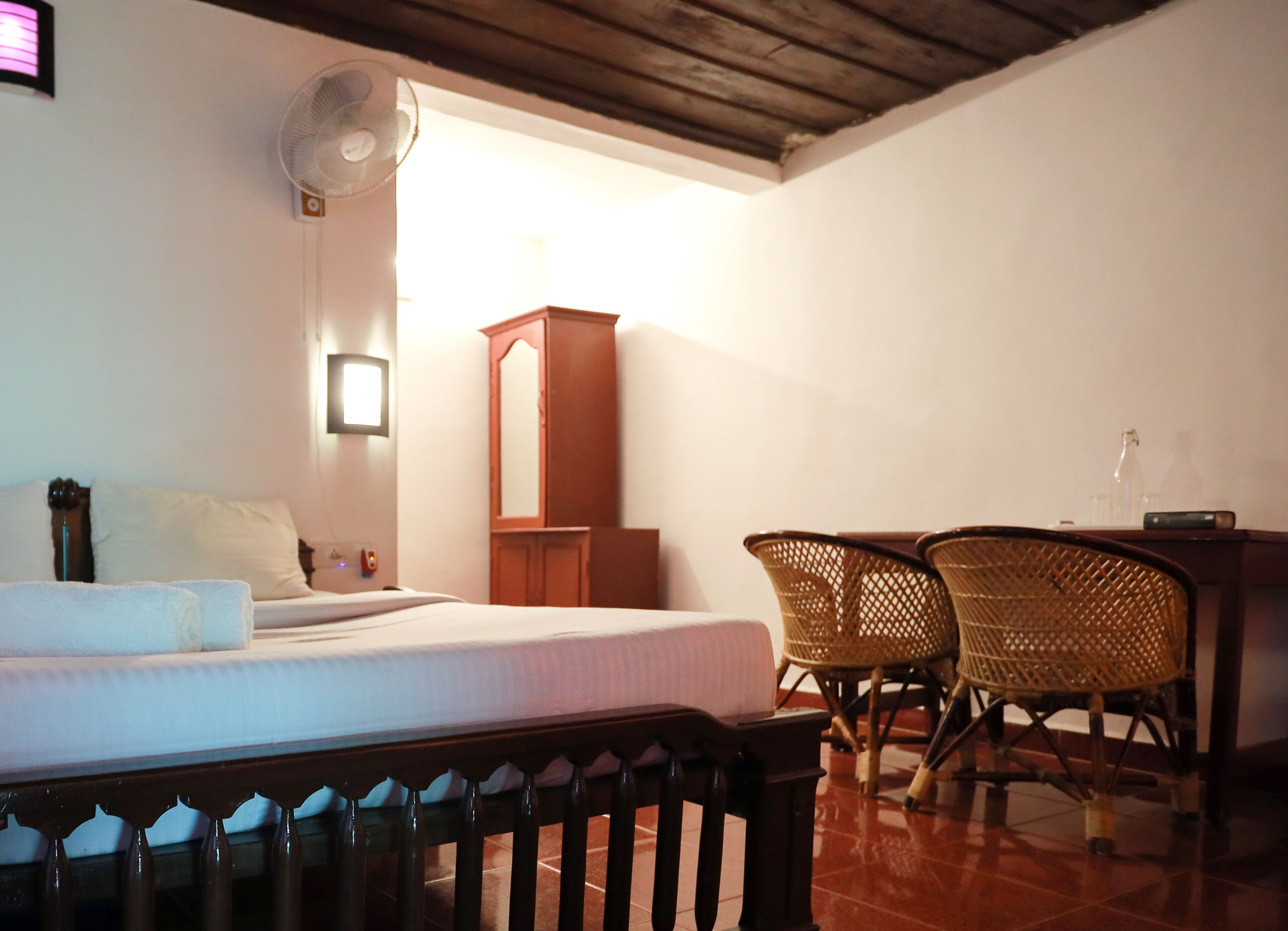 A room with a bed, a wooden cabinet, two wicker chairs, a table, and a standing fan.