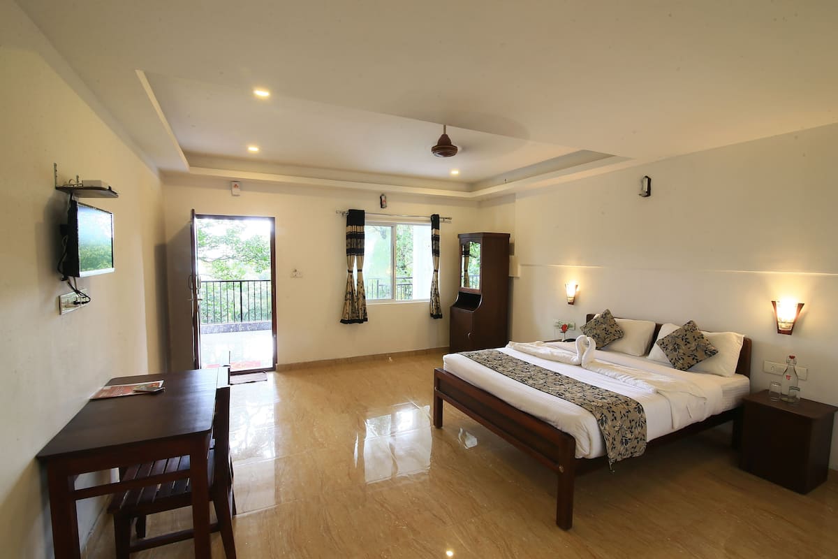 A spacious room with a bed, a desk, and a chair. There is a window with curtains and a view of greenery outside.