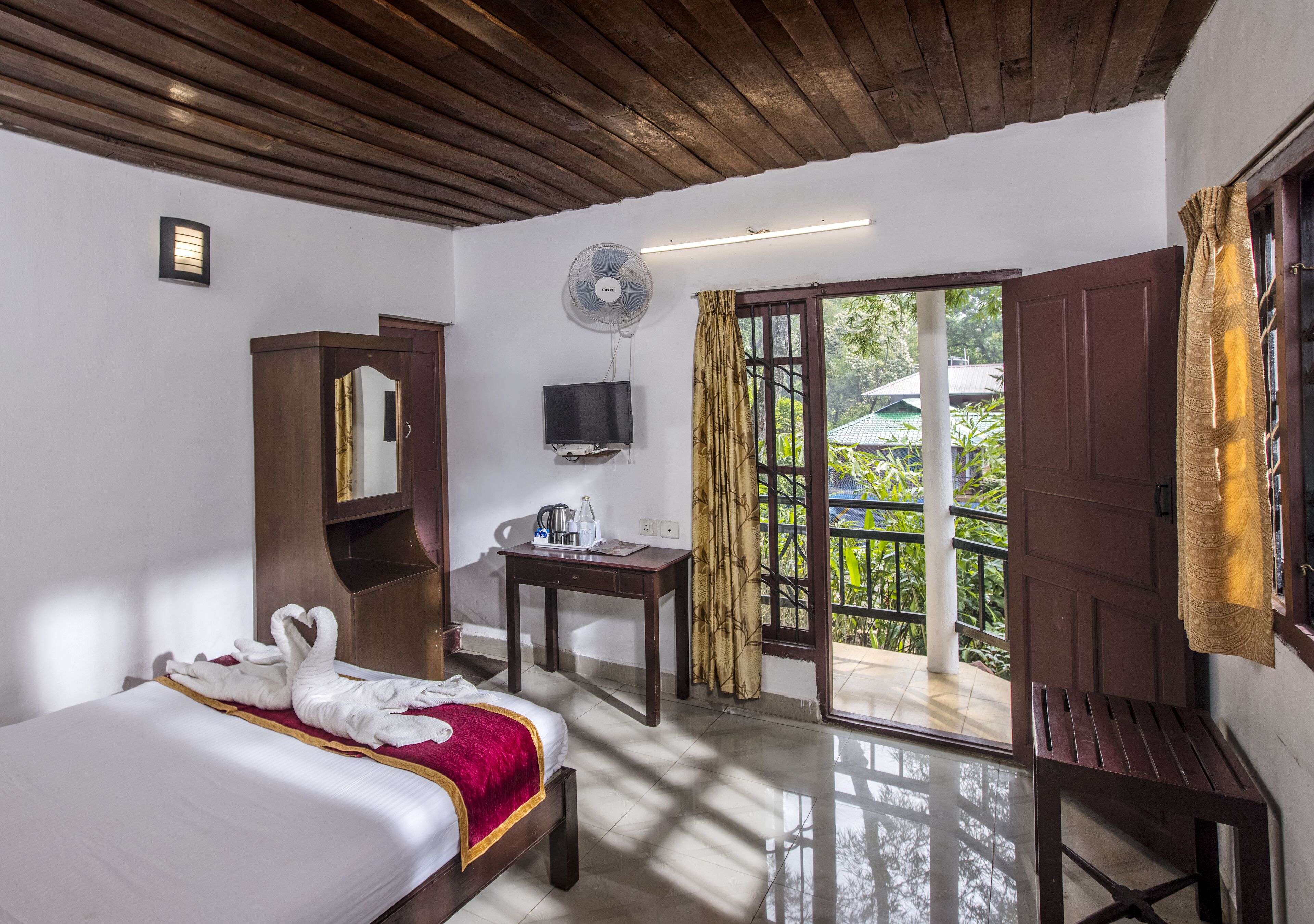 Une chambre d’hôtel avec un lit, un bureau, une télévision, un ventilateur et un balcon avec des rideaux.