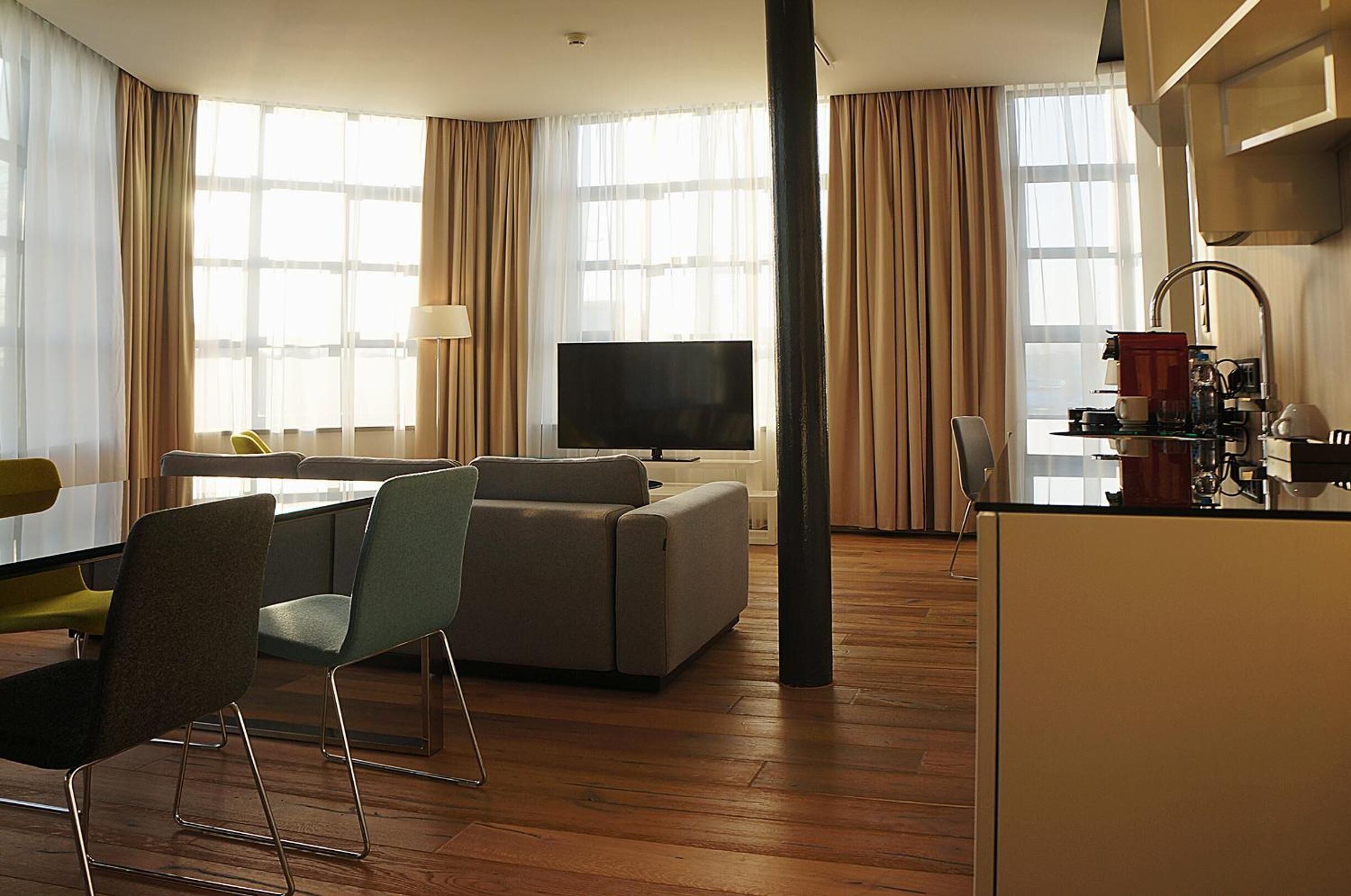 A modern hotel room with a sofa, chairs, a TV, a coffee machine, and a dining area.