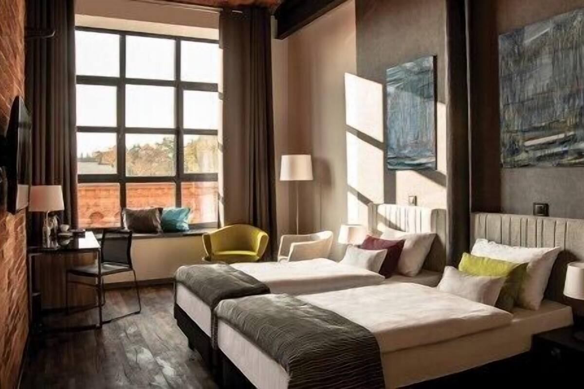 A hotel room with a brick wall, a large window with curtains, a desk with a lamp, two beds with pillows, and a chair.