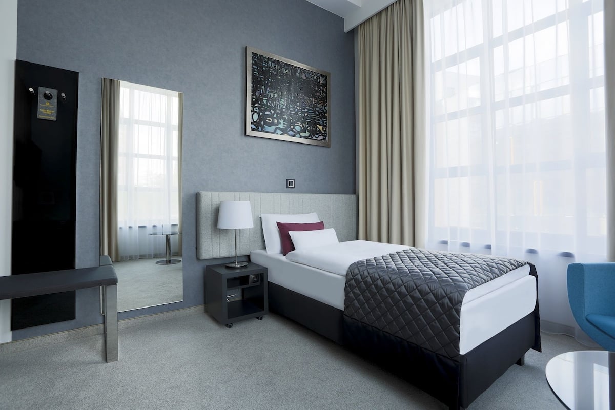 A modern hotel room with a large bed, a bedside table with a lamp, a chair, a large window with curtains, and a framed artwork on the wall.