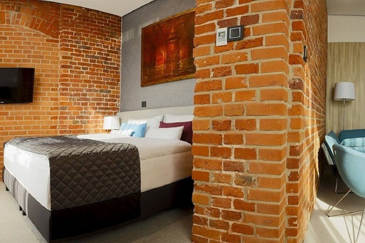 A hotel room with a brick wall, a bed with a quilted cover, a blue armchair, and a flat-screen TV mounted on the wall.
