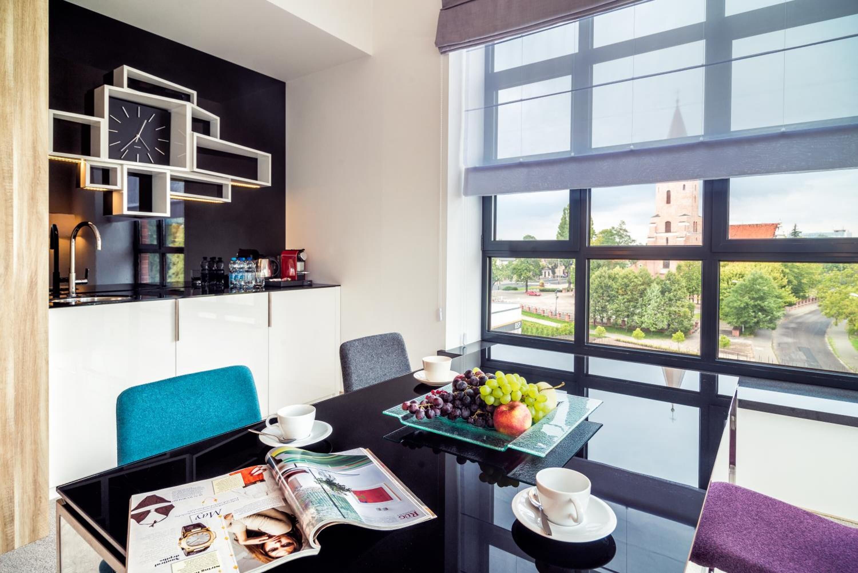 A modern room with a dining table, chairs, a kitchen area with a clock, and a large window offering a view of a building and greenery.