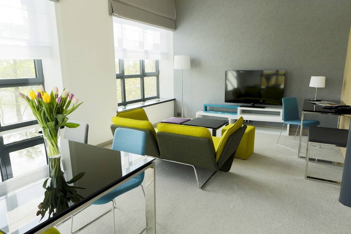 A modern hotel room with a glass table, colorful chairs, a flat-screen TV, and a vase of flowers.