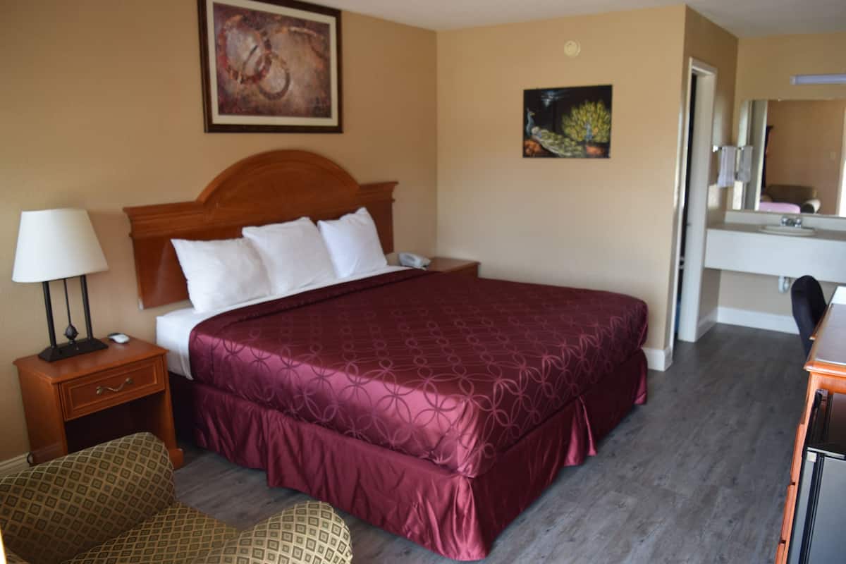A hotel room with a large bed, a bedside table with a lamp, a chair, a desk with a mirror, and a bathroom visible through an open door.