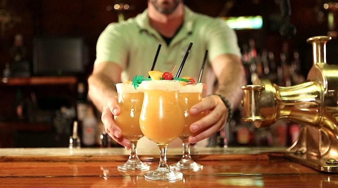 A bartender holding two cocktails with garnishes, a gold-colored bar tap, and a blurred background with a television.