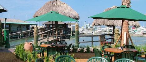 An outdoor seating area with green chairs and tables, thatched umbrellas, and a view of a marina with boats.