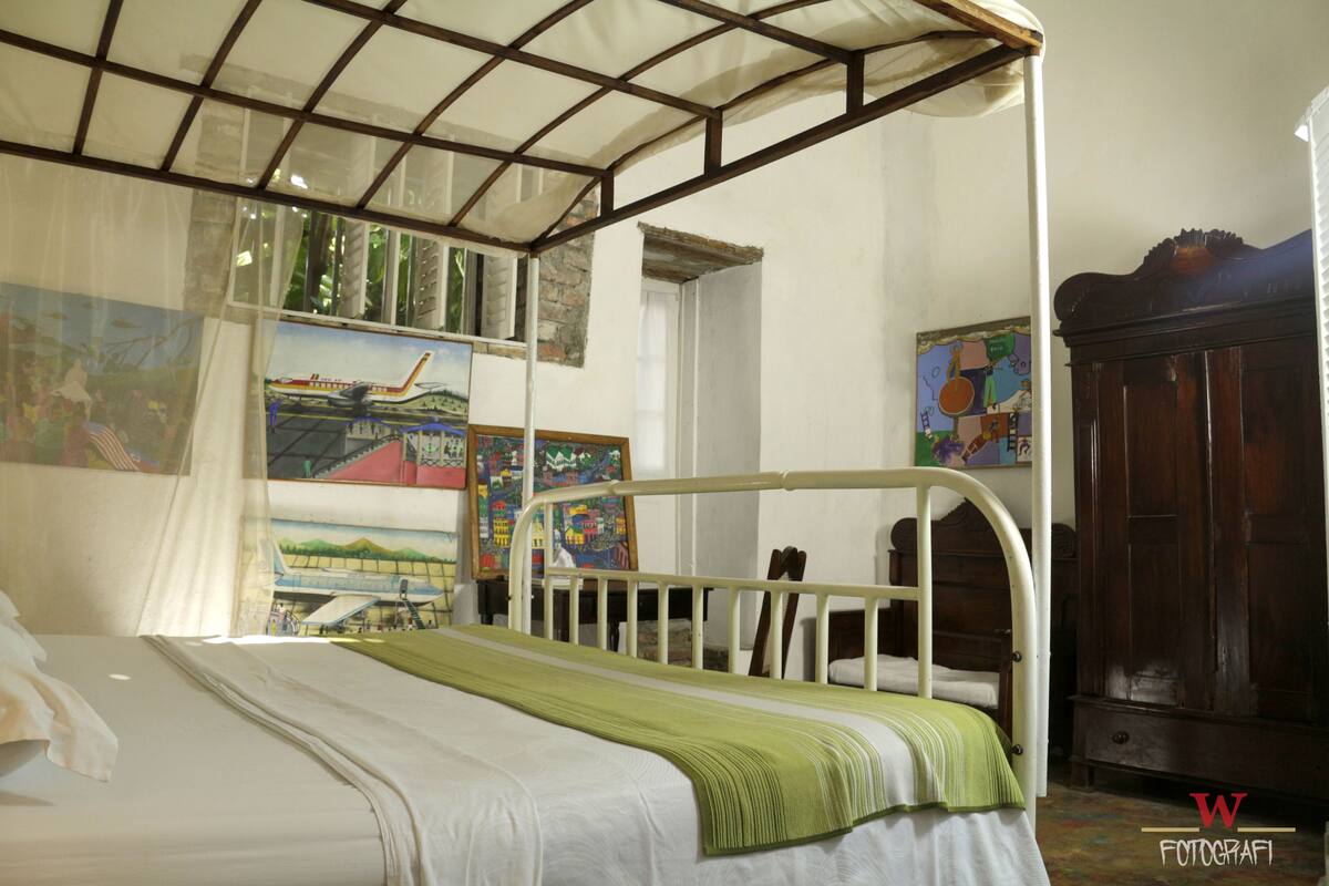 A room with a bed, a wooden wardrobe, and various paintings on the walls.