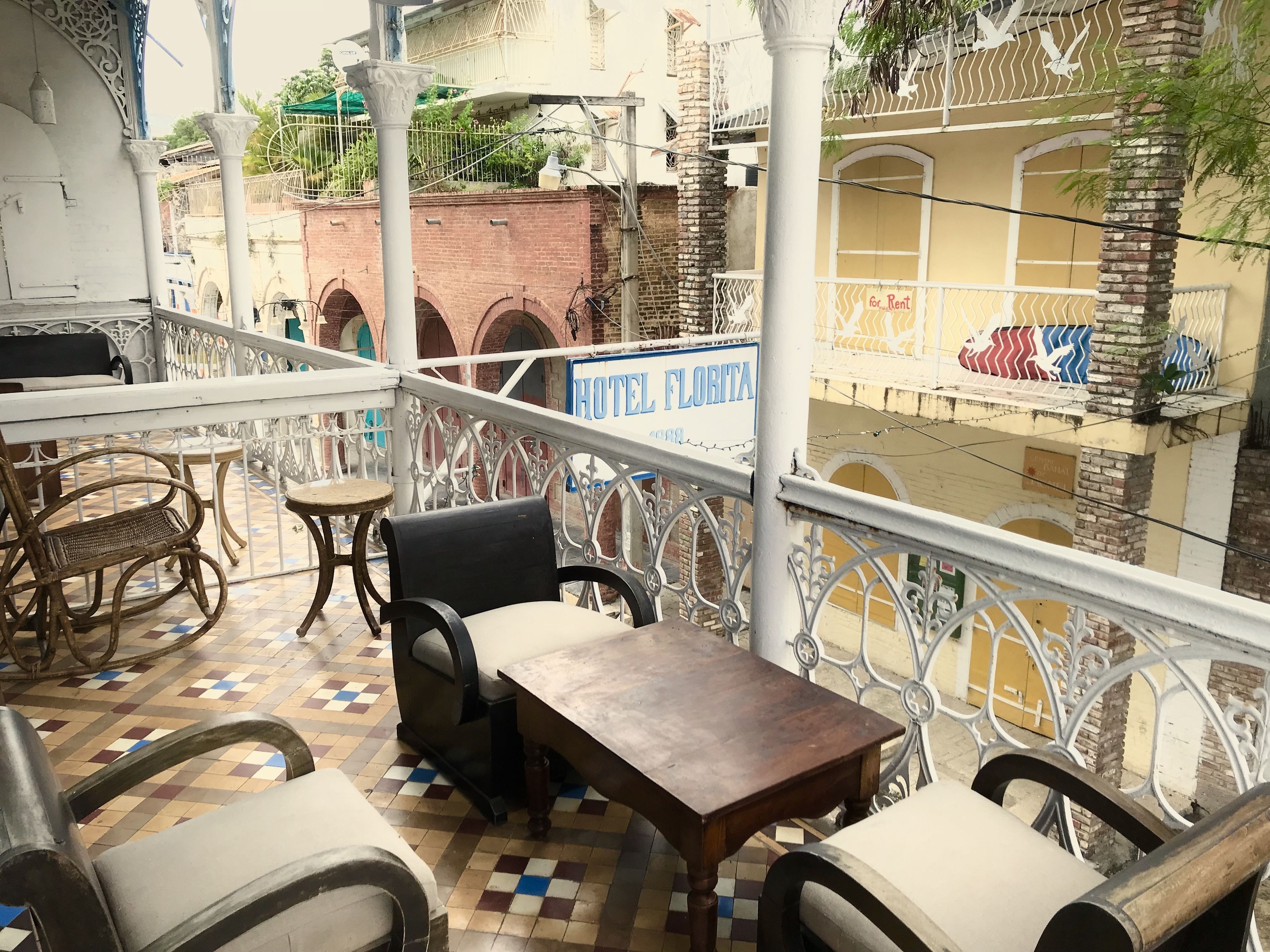 A balcony with wicker chairs and a wooden table, overlooking a street with a building labeled "HOTEL FLORITA".