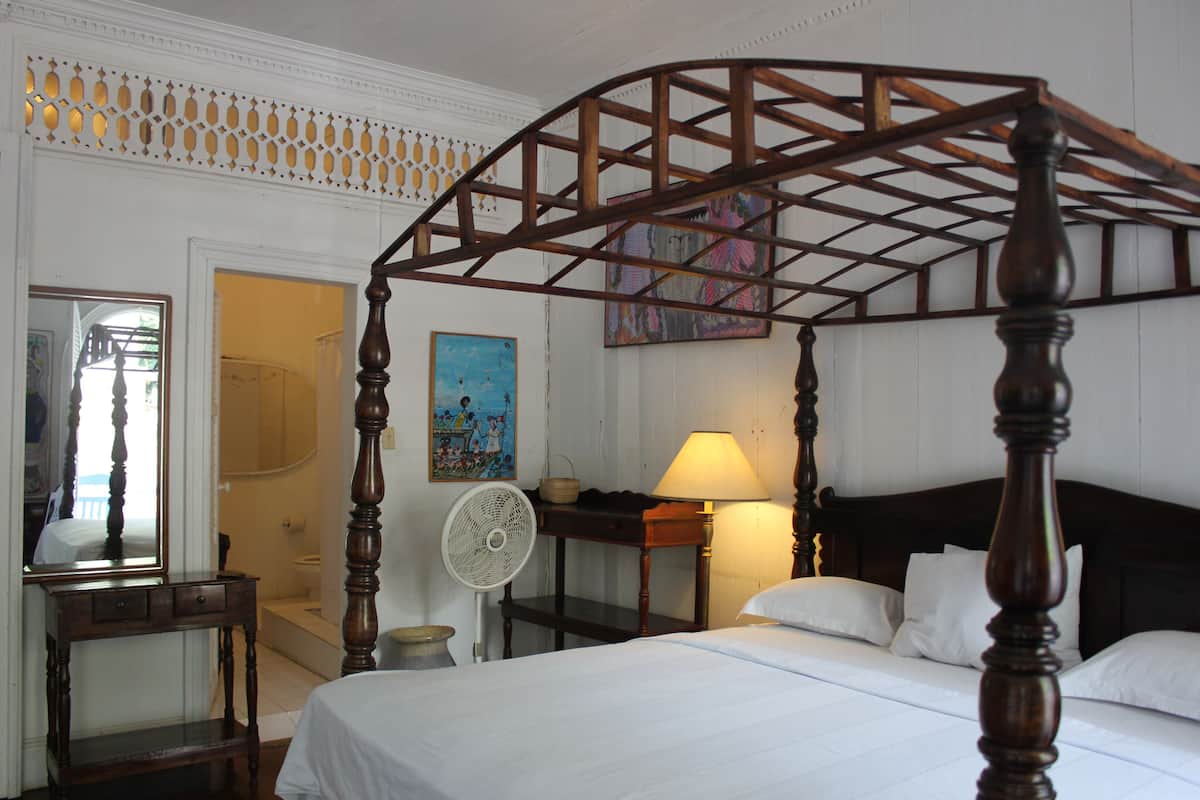 A four-poster bed with a wooden frame, a bedside table with a lamp, a fan, and a painting on the wall.