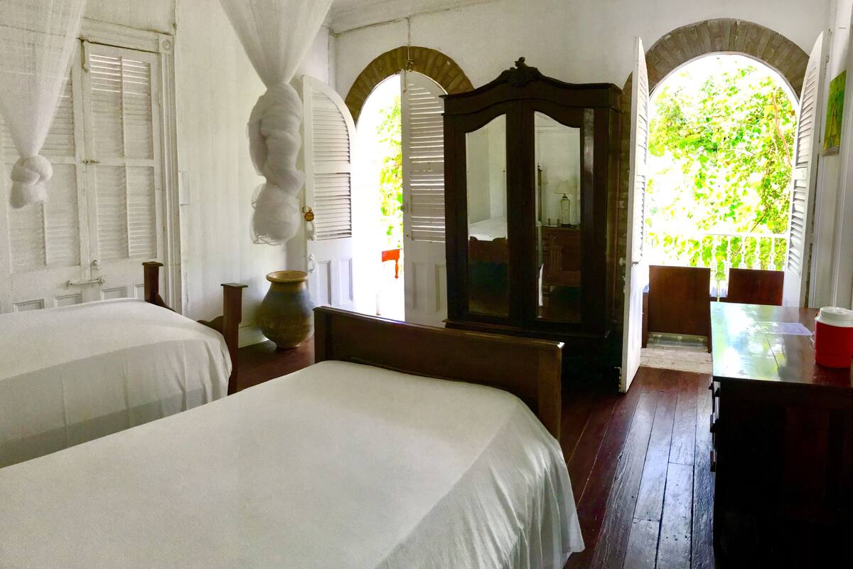 A room with two beds, a large mirror, a wooden table, and a view of greenery through an arched window.