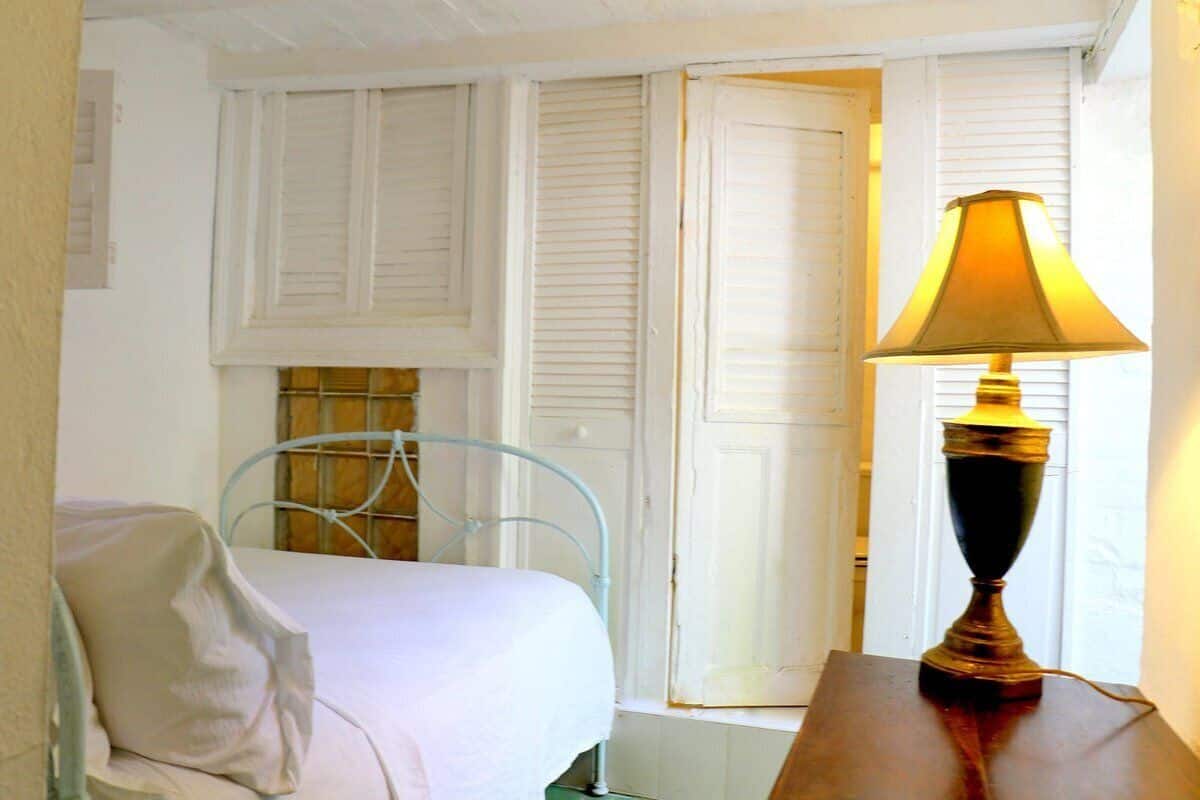 A bedroom with a bed, a metal chair, a table with a lamp, and a window with shutters.