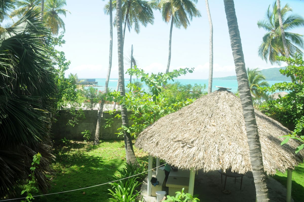 A thatched-roof hut surrounded by lush greenery and palm trees, with a view of the ocean in the background.