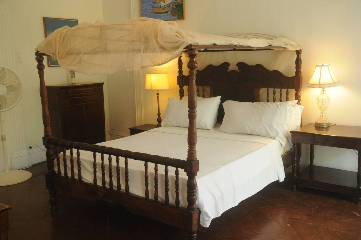 A four-poster bed with a canopy, a bedside table with a lamp, a nightstand, a dresser, and a wall-mounted fan.