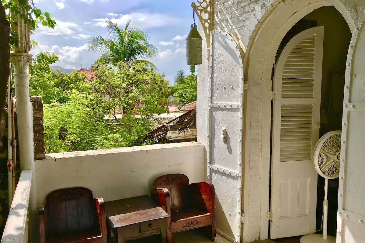 A balcony with two wooden chairs and a small table, overlooking a lush green landscape with trees and a building in the background.