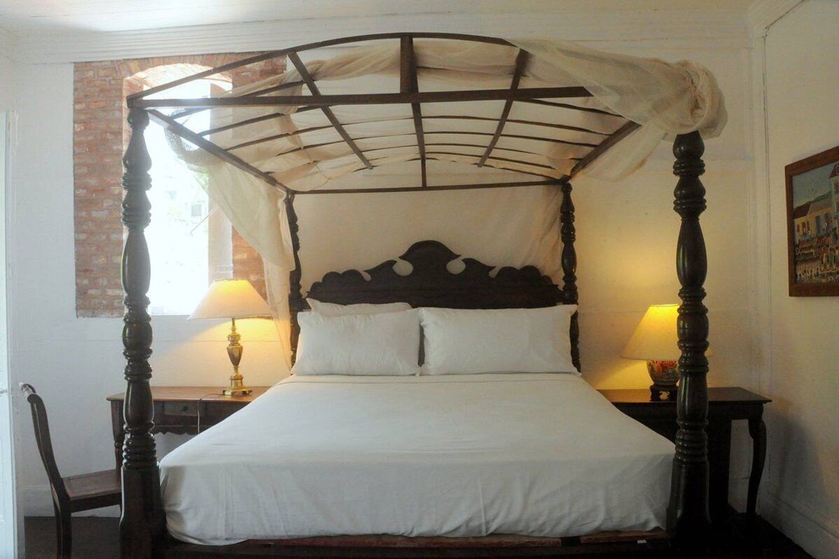 A four-poster canopy bed with white linens, two bedside tables with lamps, and a brick wall to the left.