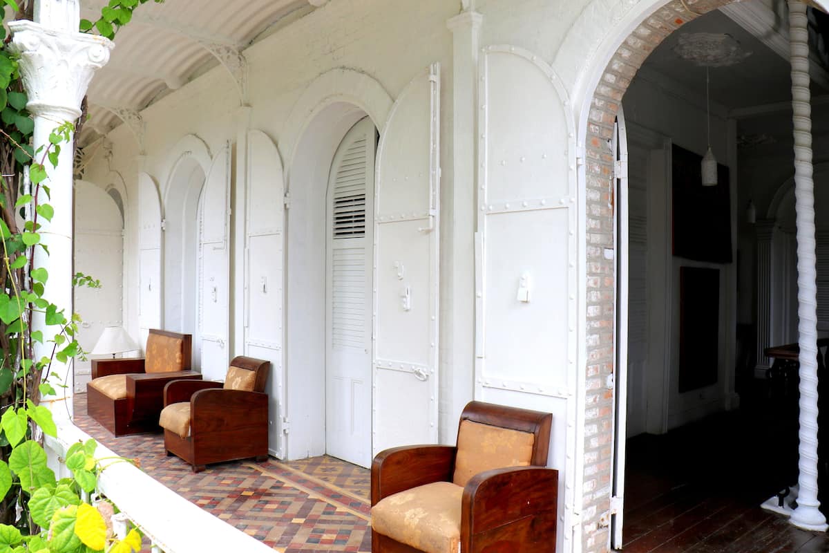 A covered porch with white columns and arches, wooden chairs, and a patterned floor.